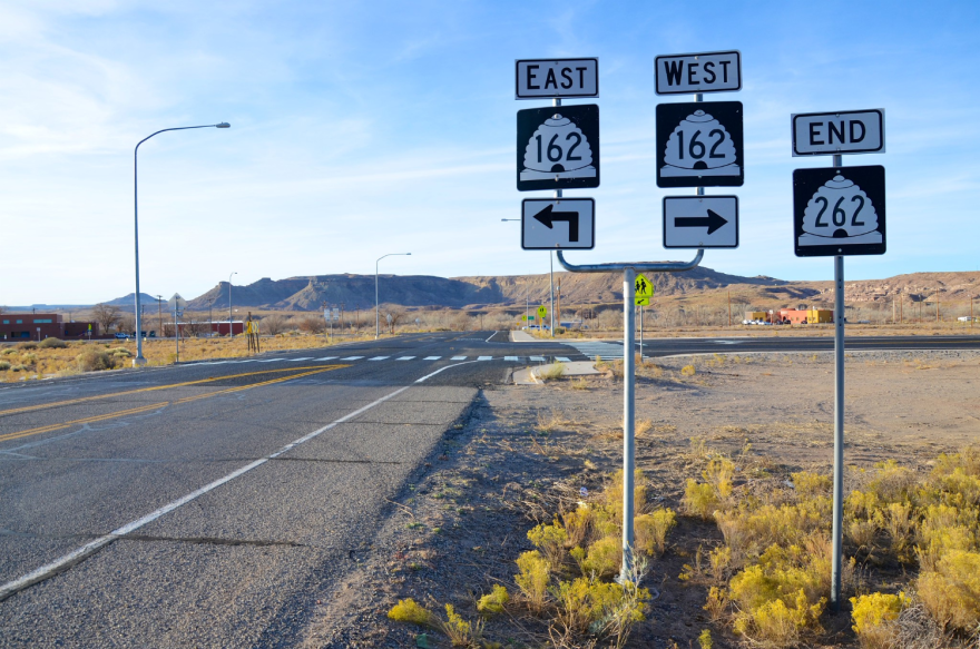 Road signs off a rural street in an arid area. There are a few buildings in the distance and mountains beyond that.