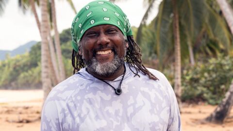 African American man with greyish beard and brown dreads wears a bandana on beach smiles into camera 