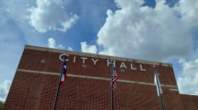A copper color brick building with white big letters that say City Hall. The Texas flag, U.S. flag and city flag is in front of the building. 