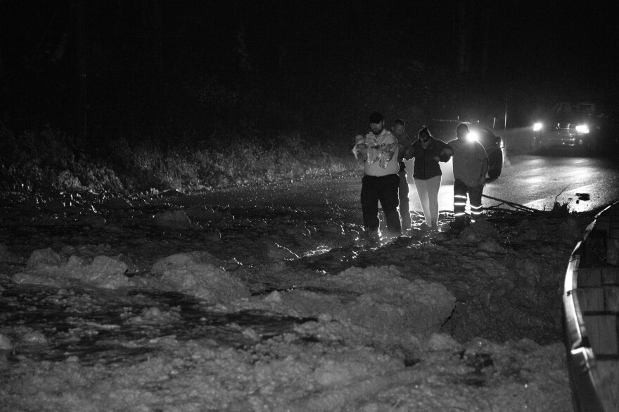 A paramedic carries a child over a mudslide to an ambulance waiting on the other side. Two other rescuers help the child's mother through the foot-deep debris.