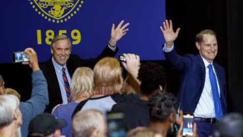 Two men in suits, standing on a stage in front of a blue Oregon state flag with "1859" written on it, wave to a crowd of people taking photos with their phones. The man on the left wears a black suit with a red-and-blue striped tie; the man on the right wears a blue suit and tie. Both are smiling.