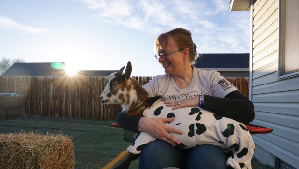 A woman holds a miniature goat dressed in a white onesie with hearts on it.