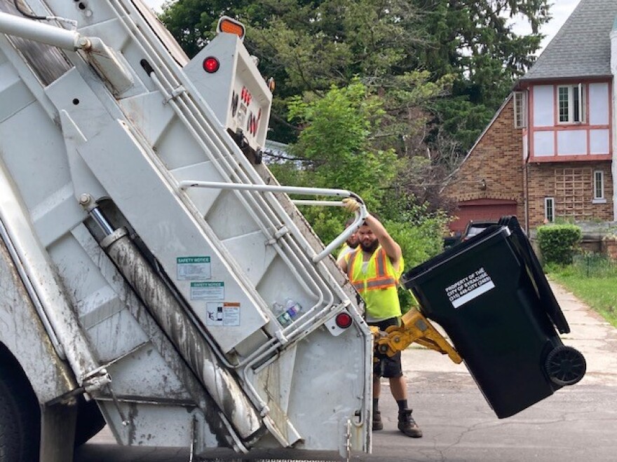 A man stands at the back of a trash truck as a mechanical arm lifts a black trash can.