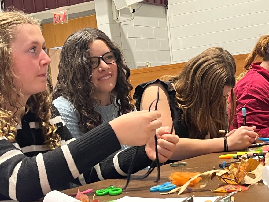 Three high school students are sitting next to each other at a table. One with blonde curly hair, is holding a piece of yarn and facing forwards. Another girl, with long dark brown curly hair and glasses, in smiling at something in another direction. Another girl with brown hair has her face down as she is coloring with a blue marker.