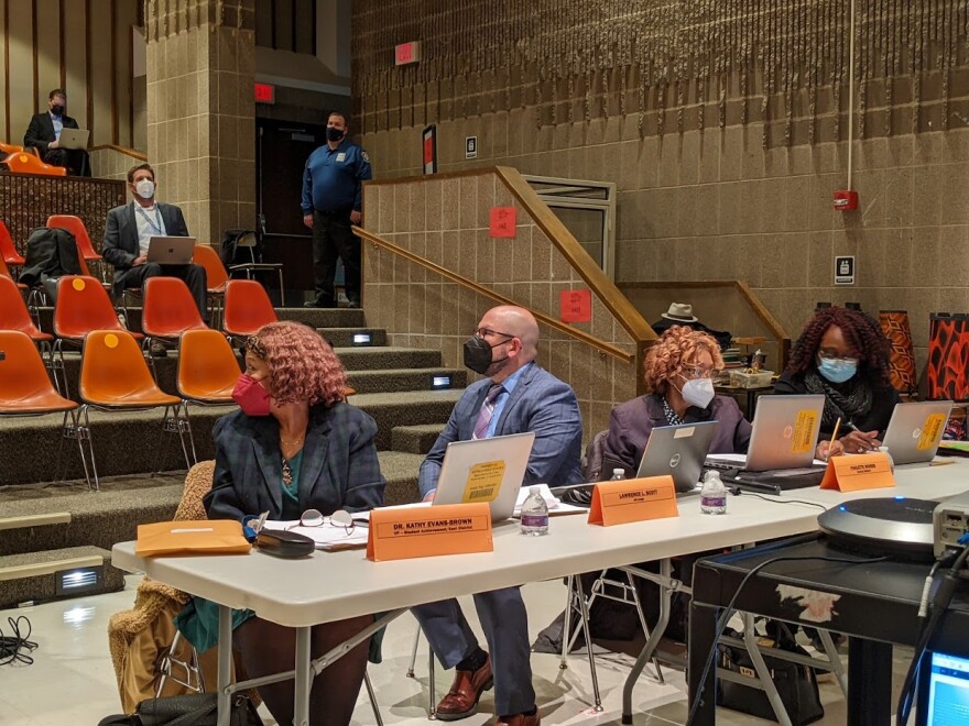 Buffalo School Board members sit at a table with their laptops, in front of an auditorium of orange stadium eating.