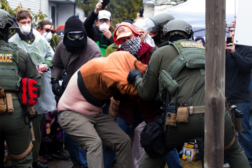 A Vermont State Police officers pulls a man from the stoop of a home where they planned to carry out an arrest, pulling up his sweater, as other protesters look on.