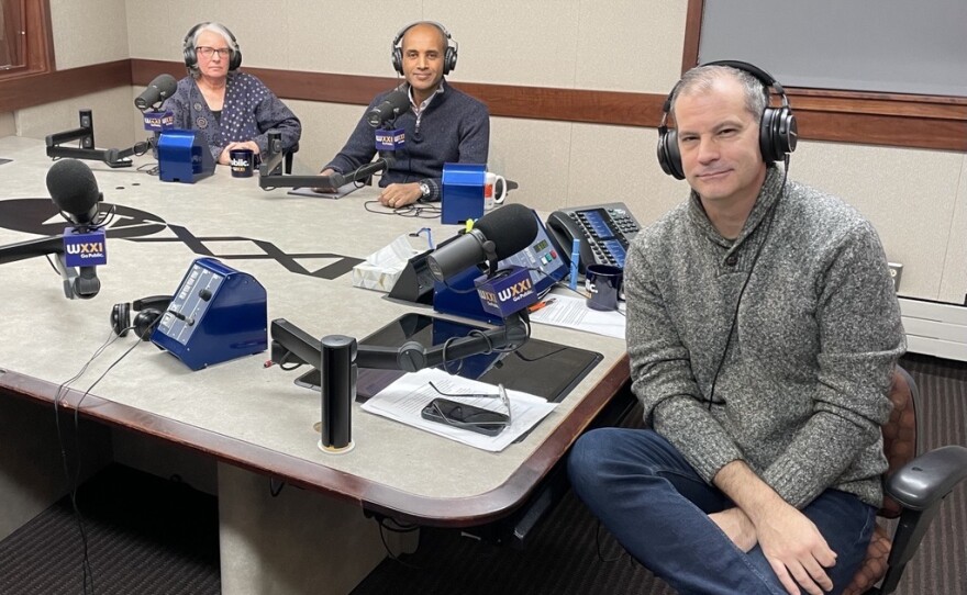 Three people wearing headphones sit at a table in a radio talk studio: a woman at left has short grey hair and is wearing glasses and a blue and white patterned shirt; a man at center is bald and is wearing a blue sweater; a man at right has short dark hair and is wearing a grey sweater, jeans and sneakers.