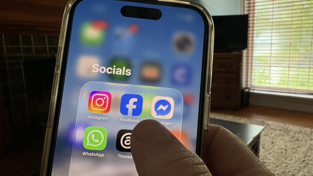 Close-up of a hand holding an iPhone. The screen reads “Socials” and in a collection are icons for Instagram, Facebook, Messenger, WhatsApp and Threads, all apps controlled by Meta.