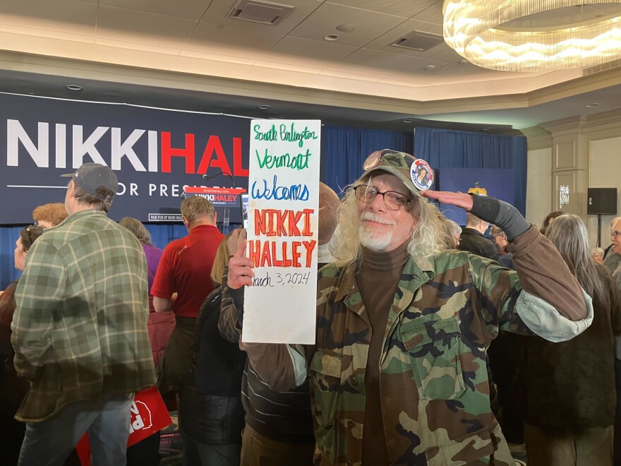 Man in cameo jacket wearing a hat holds a sign and salutes. 