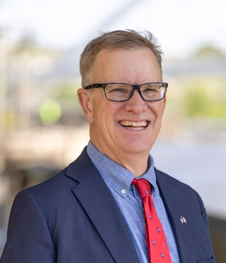 A headshot of a man who is wearing a blue suit, with a lighter blue shirt and red tie. He has glasses and a friendly smile. He is about 60 years old.