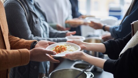 Side view closeup of unrecognizable volunteer giving hot meals to refugees in line at help center with focus on hands holding plate of rice