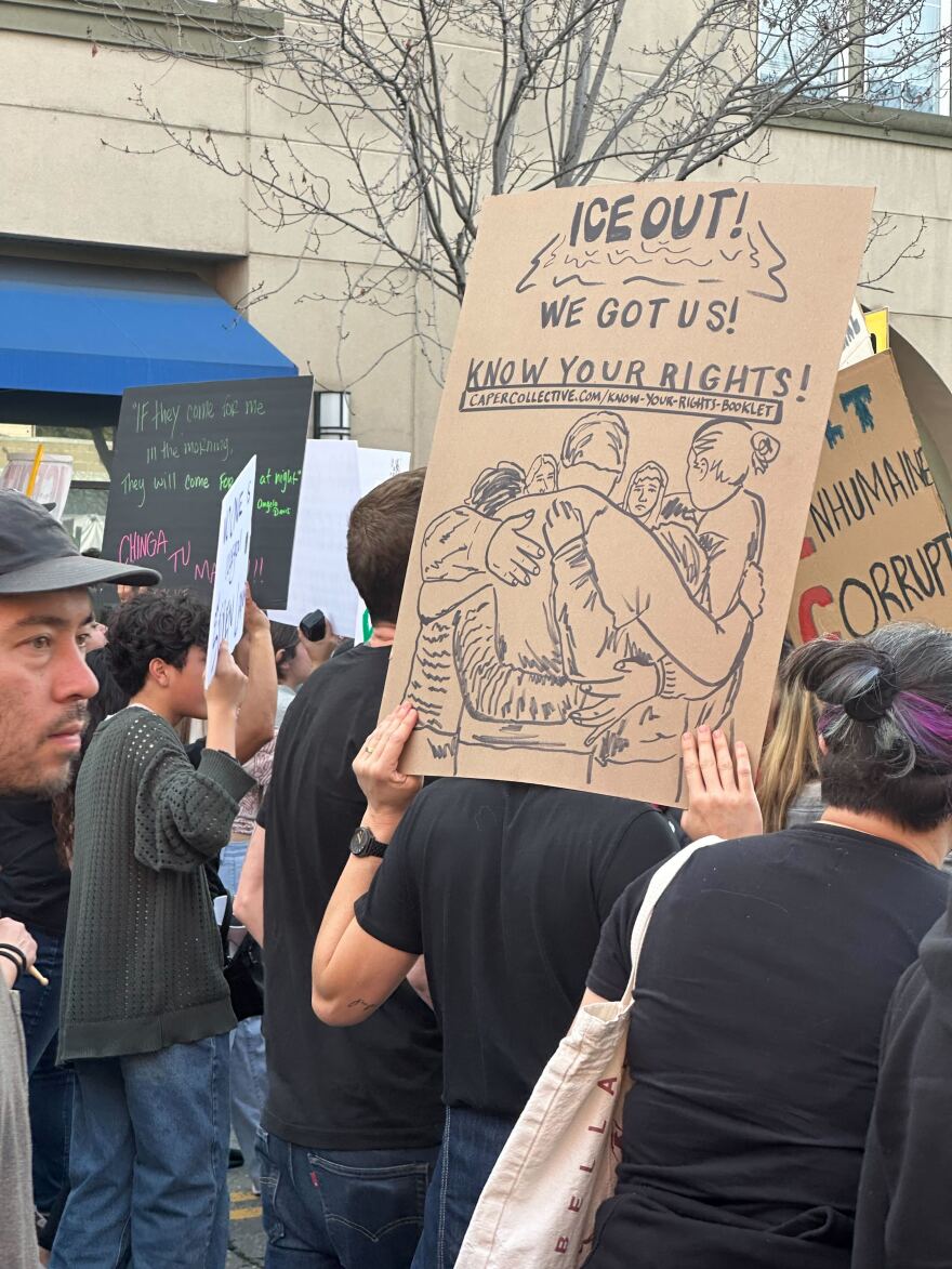 A person in a black tshirt, standing in a crowd, holds up a cardboard sign with an illstruation of people huddled together with their arms over each other's shoulders. The text reads: ICE OUT! WE GOT US! KNOW YOUR RIGHTS! CAPERCARECOLLECTIVE.COM/KNOW-YOUR-RIGHTS-BOOKLET