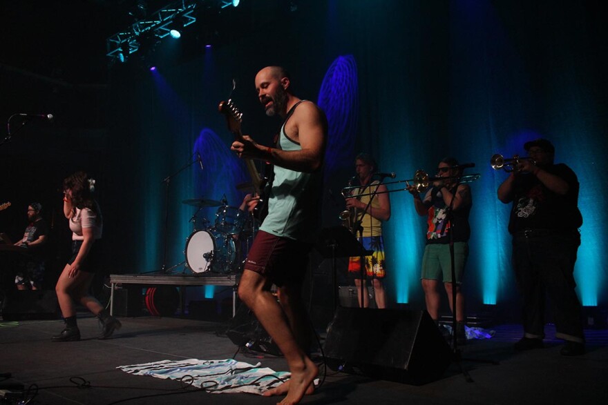 Man in shorts and tank top playing guitar with three horn players in the background