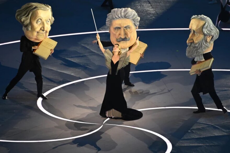 A woman in a black dress conducts three dancers dressed as famous Italian composers: Rossini, Puccini, and Verdi.