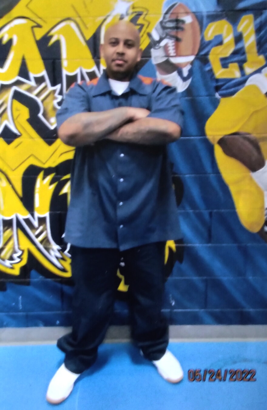A full-length photo shows a man standing with his arms crossed in front of a brightly painted mural.