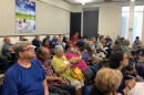  Around 20 or so people sit in rows of chairs, listening attentively. A BPU poster hangs behind them. 