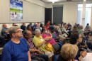  Around 20 or so people sit in rows of chairs, listening attentively. A BPU poster hangs behind them. 