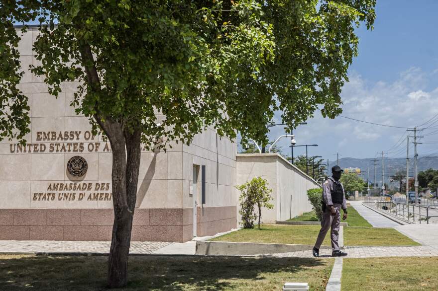 A man wearing a vest and baseball cap stands outside of the American embassy in Port-au-Prince, Haiti, against a backdrop of mountains.