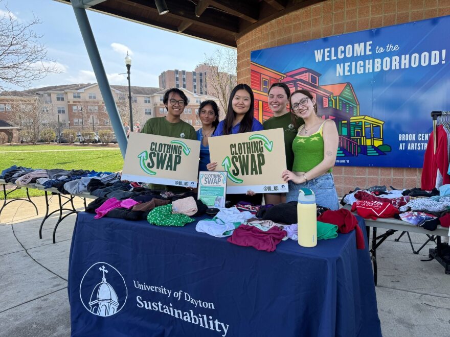A group of five students stand behind a table covered in stacks of clothes. They hold signs that say, 'Clothing Swap.'