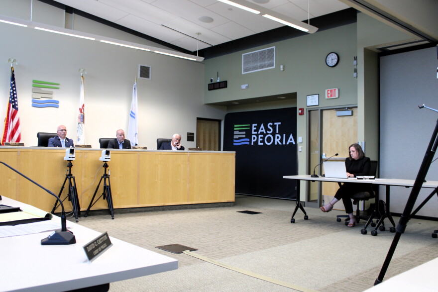 Three officials sit at a dais in a city council chamber, while a woman works on a laptop at a separate table in front of a wall sign reading “East Peoria.”