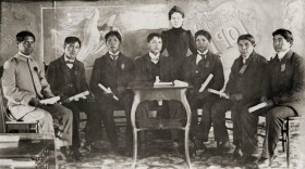 A photo from 1901 of a group of seven Native American male students sitting in chairs, facing the camera. None of them are smiling. A white female teacher is standing behind them.