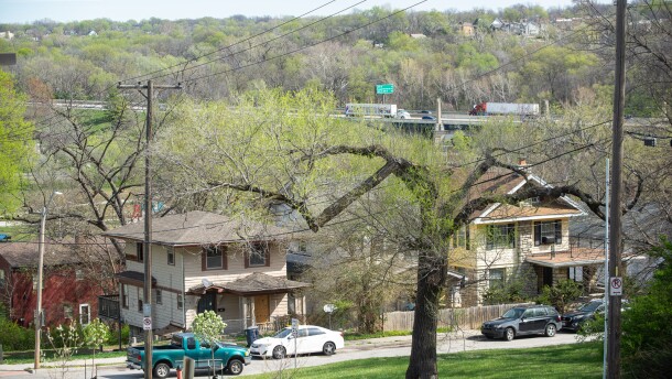 A pair of two-story houses sit in the foreground with cars parked in the street. In far background, a highway can be seen with traffic moving along it.