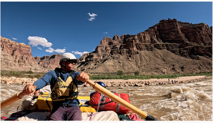A man pushes the oars of his boat against tough rapids. His lips are pursed tightly as he has to push hard against the rapids. He has various bags behind him and is wearing a yellow lifejacket and white helmet.