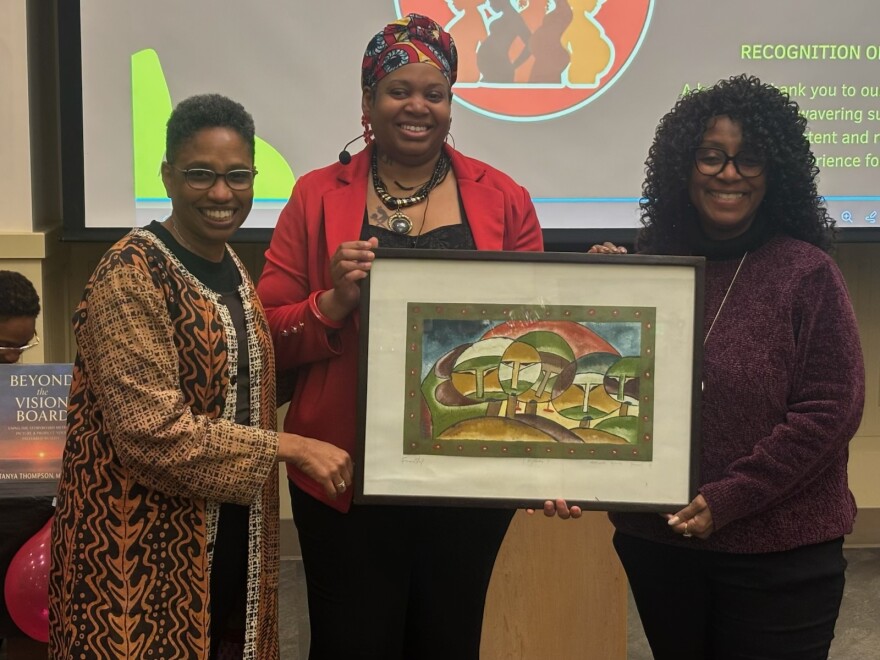 Three women holding a framed work of art pose with a screen projecting computer slides behind them.