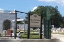 A sign that reads MARTI COLON CEMETERY/3110 WEST COLUMBUS DRIVE/TAMPA PARKS AND RECREATION DEPARTMENT stands in front of an open gate. Headstones and mausoleums are visible behind on the cemetery lawn. An older couple is visible in the left foreground, walking outside the entrance.