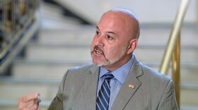 A bald white man with a short beard and wearing a gray suit with blue shirt and striped tie speaks in front of a set of stairs.