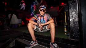 A man in sunglasses and colorful clothing sits on a stage while smiling for the camera