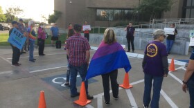 IMG_8399.JPG Woman at a distance addresses a crowd of a few dozen facing her as she holds a microphone outside, as the sun is going down.