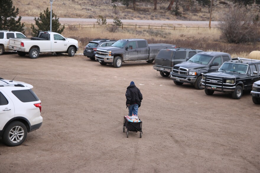 A man in a black hoodie wheels a canvas-sided wagon full of groceries out to a gravel parking lot.
