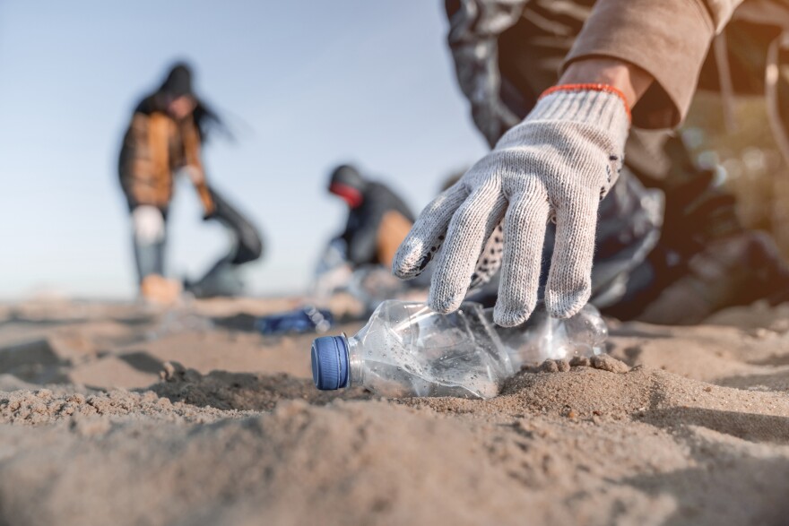 A gloved hand picks up a discarded plastic bottle on a sandy beach, with other volunteers blurred in the background during a cleanup