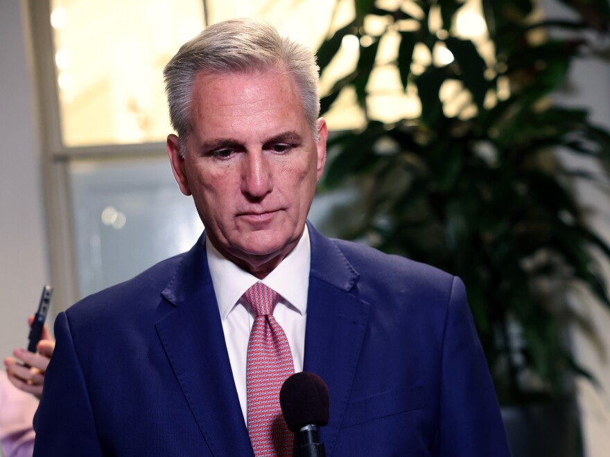 A close-up of a man looking forward and downward. There is a microphone pointed toward him in the foreground.