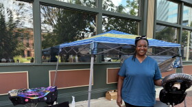 Woman in blue top and dark pants smiles for a portrait while standing under a pop-up awning, next to a massage chair