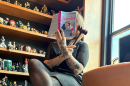 A reader covers her face from view of the lens with an open copy of Overruled by Lana Ferguson in her left hand, her right hand holding a gavel. She's seated on a desk near an open window, a bookshelf completely stocked with fantasy character toys behind her.