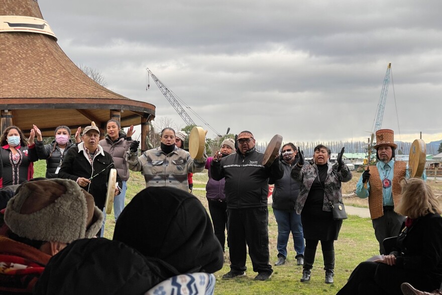 Four people playing hand drums are surrounded by others with hands raised on a grassy berm. There is water and a cedar pavilion in the background and people sitting in front of the performers. 