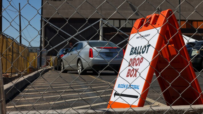 Orange, white and blue sign with arrow pointing to Ballot Drop Box seen through fence with cars behind it
