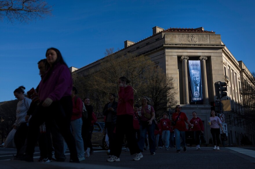 Visitors cross Pennsylvania Avenue NW near a banner of President Donald Trump, outside the U.S. Department of Justice on Saturday, March 21, 2026, in Washington.