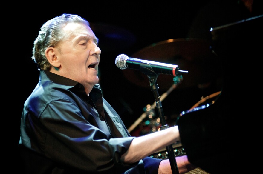 An older white man with gray hair combed back, wearing a black shirt sings into the mic while playing the piano.