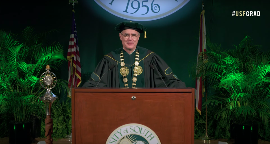 man in graduation cap and gown speaks at podium