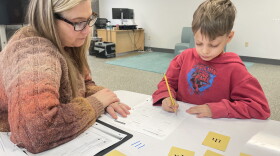 An adult in a sweater and glasses watches a child in a red hoodie complete a worksheet. 