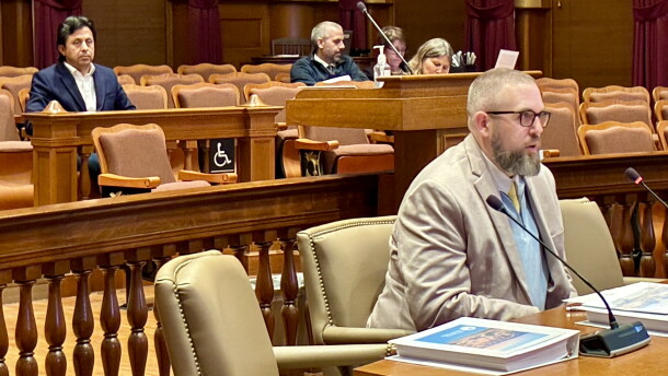 A man with a beard and glasses sits at a table in a courtroom, with other people seated in the background.