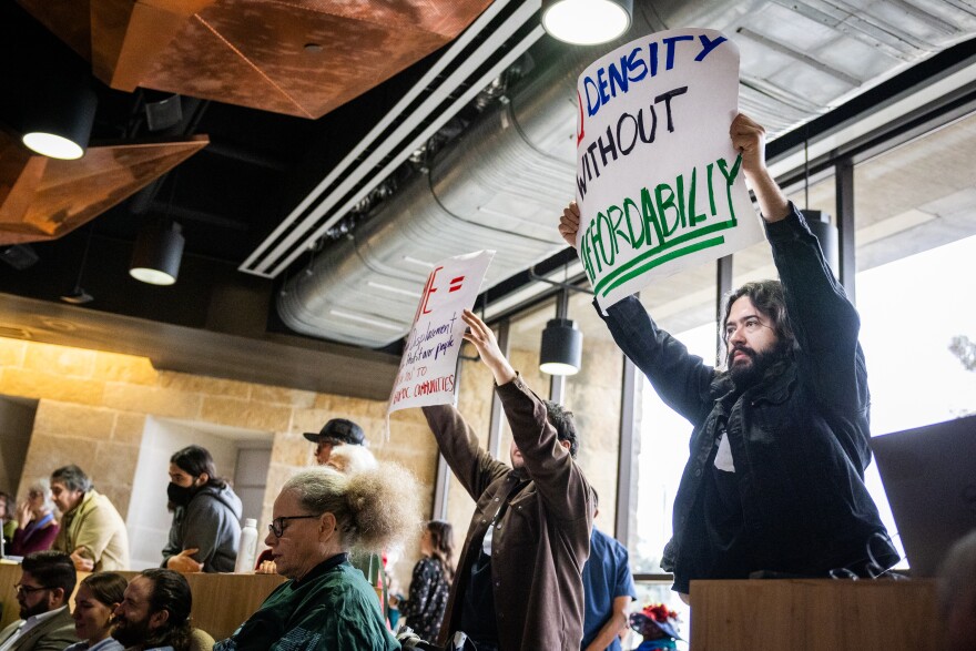 People hold signs that say "No density without affordability" at the back of a room with others seated in front of them