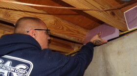 An over-the-shoulder image of a bald man in a navy blue hoodie placing a piece of purple foam insulation in between the beams of a ceiling. 