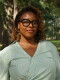 A smiling woman with black glasses stands in front of trees and shrubbery.