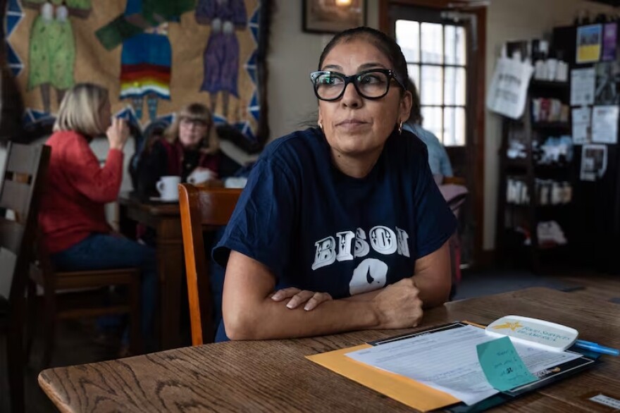 A woman sits at a table in a coffeeshop, with paperwork in front of her. 