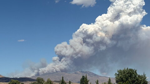 Smoke fills an otherwise clear, blue sky from behind rolling hills.