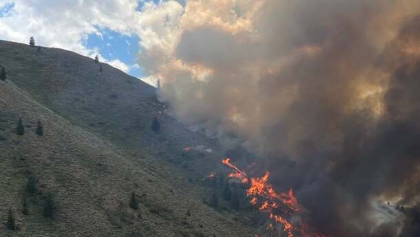 A brushfire burns in the hills just outside Hailey, ID, with bright orange flames and dense black smoke rising over a mountain. 